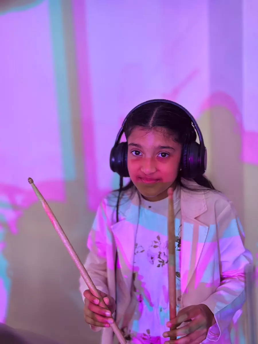 A young girl wearing headphones and holding drumsticks against a vibrant background
